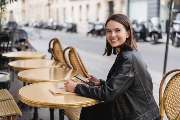 Cheerful woman in leather jacket holding smartphone near notebook on round table in outdoor cafe terrace — Stock Photo