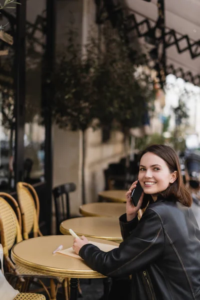 Femme gaie en veste en cuir parler sur smartphone et assis dans un café extérieur en france — Photo de stock