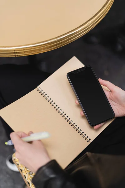 Hochwinkel-Ansicht einer Frau mit Smartphone mit leerem Bildschirm und Notizbuch in der Nähe des runden Tisches — Stockfoto