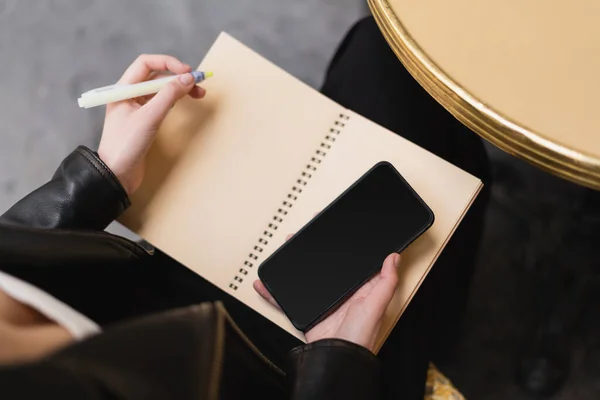 Partial view of woman holding smartphone with blank screen and notebook near round table — Stock Photo