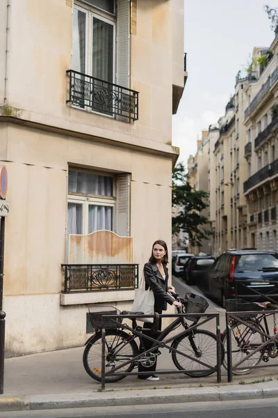 Young stylish woman in black leather jacket standing near bicycle on street in paris — Stock Photo