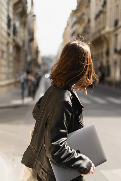 Задний вид фрилансера в черной кожаной куртке, идущего с ноутбуком на улице в Париже — стоковое фото
