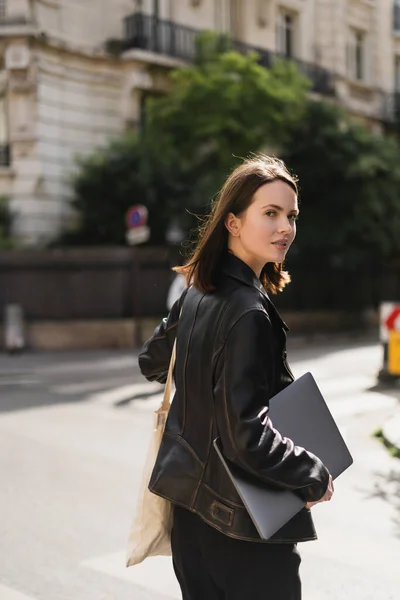 Young freelancer in black leather jacket walking with laptop on street in paris — Stock Photo