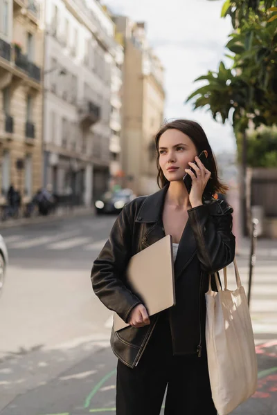 Young freelancer in black leather jacket holding laptop and talking on smartphone on street in paris — Stock Photo