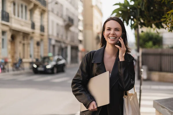 Happy freelancer in black leather jacket holding laptop and talking on smartphone on street in paris — Stock Photo