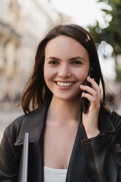 Portrait de femme joyeuse veste en cuir noir tenant ordinateur portable et parlant sur smartphone dans la rue à Paris — Photo de stock