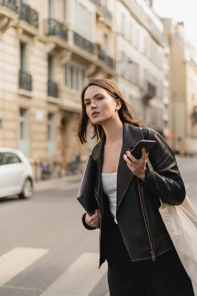 Mujer con estilo en chaqueta de cuero negro que sostiene el ordenador portátil y el teléfono inteligente en la calle en París - foto de stock