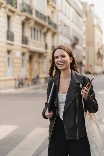 Femme gaie en veste en cuir noir tenant ordinateur portable et smartphone dans la rue à Paris — Photo de stock