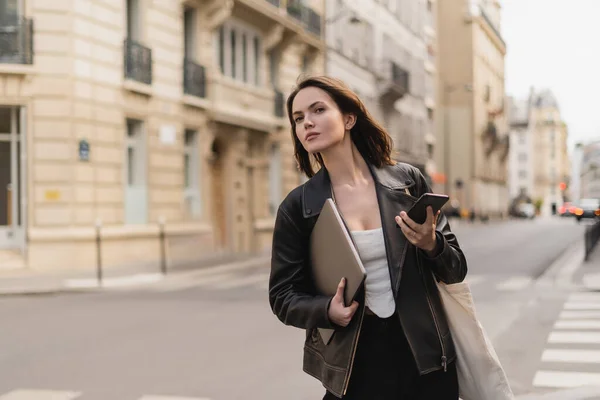 Stylish freelancer in black leather jacket holding laptop and smartphone on street in paris — Stock Photo