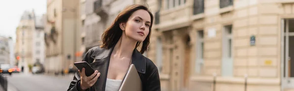 Stylish freelancer in black leather jacket holding laptop and smartphone on street in paris, banner — Stock Photo