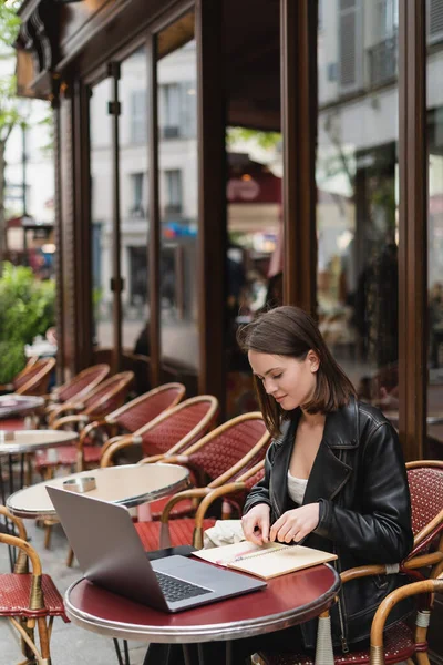 Elegante freelancer en chaqueta de cuero negro mirando el cuaderno mientras está sentado cerca de la computadora portátil en la cafetería francesa al aire libre - foto de stock