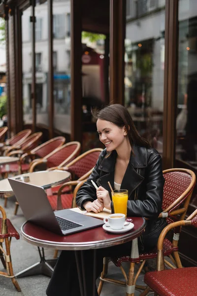 Feliz freelancer en chaqueta de cuero negro mirando portátil cerca de bebidas en la mesa en francés café al aire libre - foto de stock