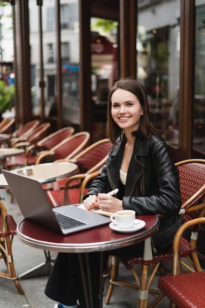 Freelancer feliz em jaqueta preta olhando para a câmera perto de laptop e xícara de café na mesa no café francês ao ar livre — Fotografia de Stock