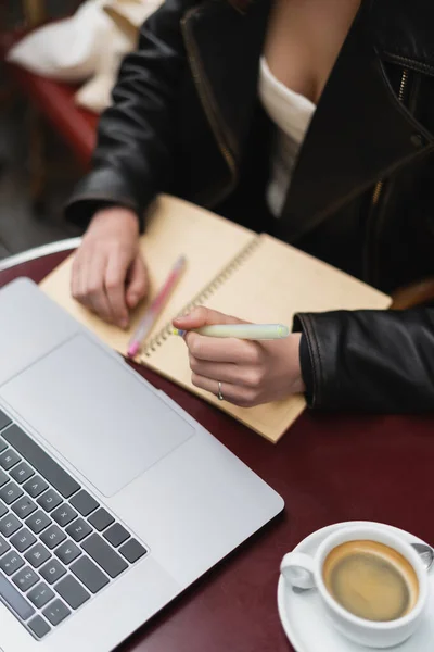 Cropped view of freelancer making notes near laptop and cup of coffee on table in french outdoor cafe — Stock Photo