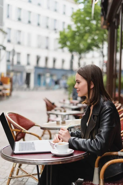 Vista lateral de sonriente freelancer en chaqueta negra mirando portátil cerca de taza de café en la mesa en francés café al aire libre - foto de stock