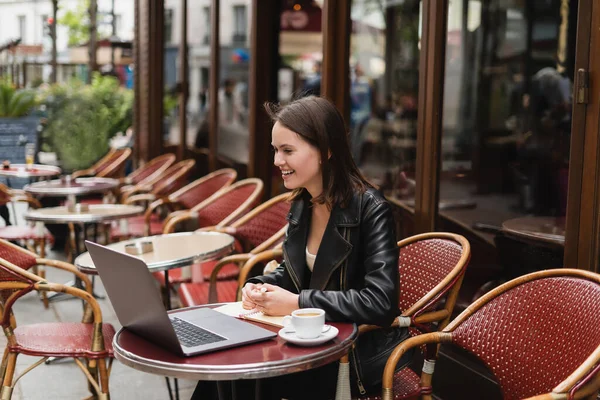 Freelance souriant en veste noire regardant ordinateur portable près de tasse de café sur la table dans un café en plein air français — Photo de stock