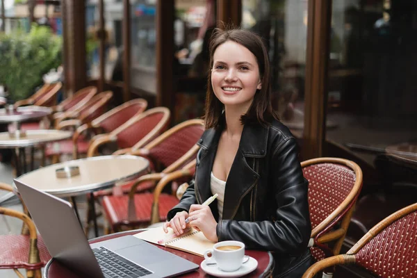 Mulher feliz em casaco preto olhando para a câmera perto do laptop e xícara de café na mesa no café francês ao ar livre — Fotografia de Stock