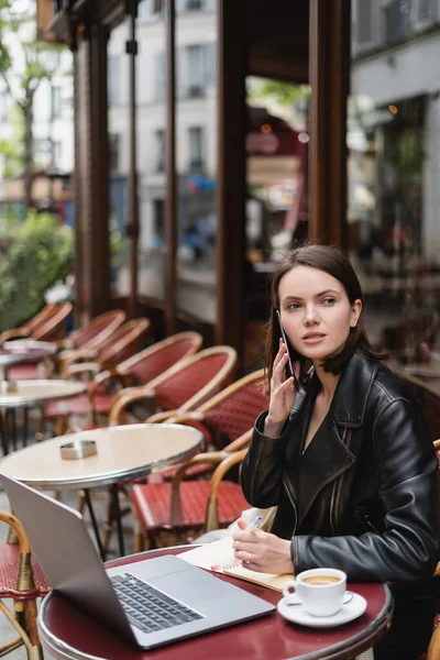 Jeune pigiste en veste noire parlant sur smartphone près d'un ordinateur portable et tasse de café sur la table dans un café en plein air français — Photo de stock