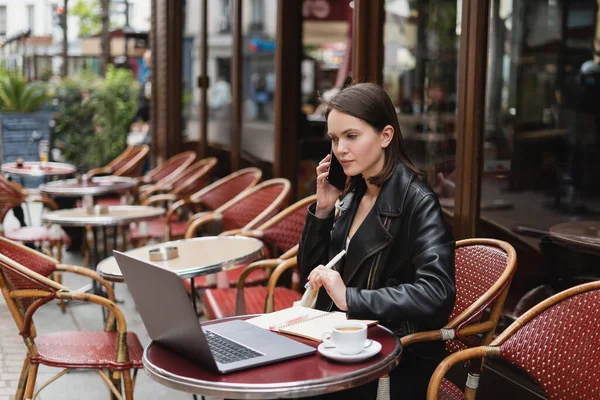 Молода жінка в чорній куртці говорить на смартфоні біля ноутбука і чашки кави на столі у французькому відкритому кафе — стокове фото