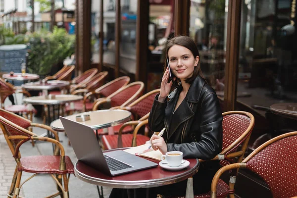 Lächelnder Freiberufler in schwarzer Jacke im Gespräch auf dem Smartphone neben Laptop und einer Tasse Kaffee auf dem Tisch in einem französischen Café im Freien — Stockfoto