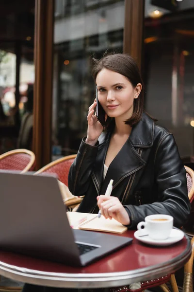 Freelancer in black jacket talking on smartphone near laptop and cup of coffee on table in french outdoor cafe — Stock Photo