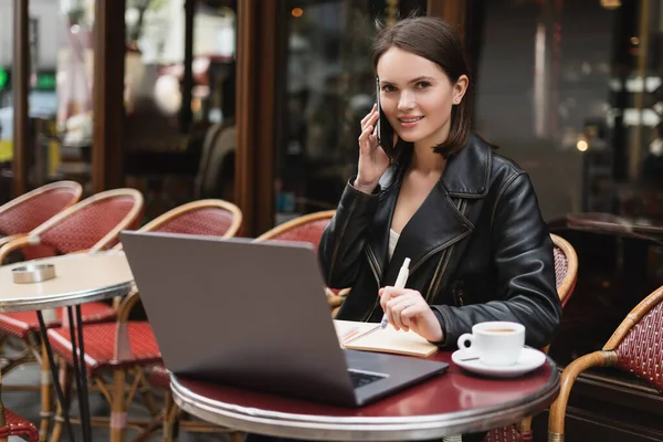 Freelance souriant en veste noire parlant sur smartphone près d'un ordinateur portable et tasse de café sur la table dans un café extérieur à Paris — Photo de stock