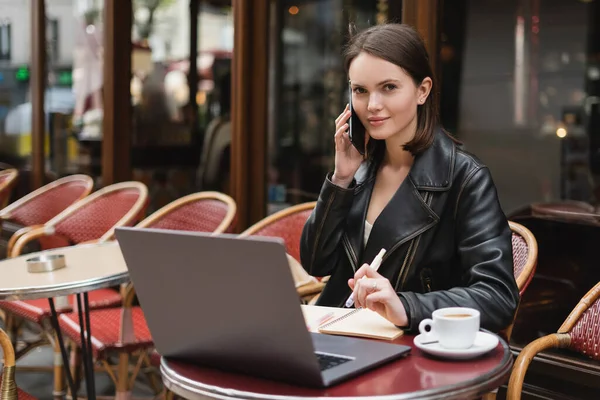 Jeune pigiste en veste noire parlant sur smartphone près d'un ordinateur portable et tasse de café sur la table dans un café en plein air à Paris — Photo de stock