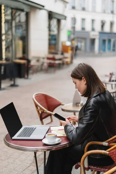 Seitenansicht von Freelancer in schwarzer Jacke mit Smartphone in der Nähe von Laptop und Tasse Kaffee in französischem Café — Stockfoto