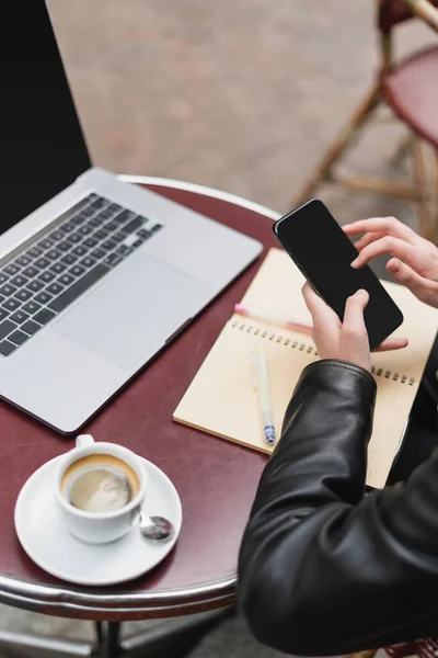 Vista cortada do freelancer em casaco preto segurando smartphone perto de laptop e xícara de café no café francês ao ar livre — Fotografia de Stock