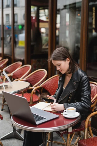 Young freelancer in black jacket using smartphone with blank screen near laptop and cup of coffee in french outdoor cafe — Stock Photo