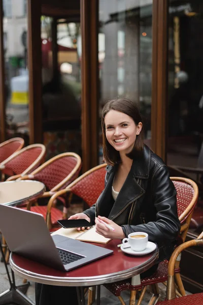 Щасливий фрілансер у чорній куртці тримає смартфон біля ноутбука та чашки кави у французькому відкритому кафе — стокове фото