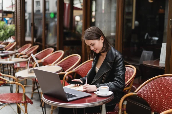 Jovem freelancer em casaco preto segurando smartphone e escrevendo perto de laptop e xícara de café no café francês ao ar livre — Fotografia de Stock