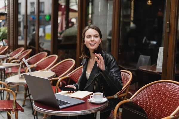 Jovem freelancer em casaco preto segurando smartphone e mão acenando perto de laptop e xícara de café no café francês ao ar livre — Fotografia de Stock