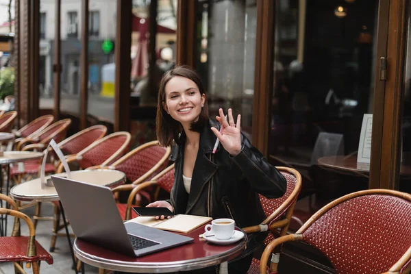 Mulher feliz em casaco preto segurando smartphone e mão acenando perto de laptop e xícara de café no café francês ao ar livre — Fotografia de Stock