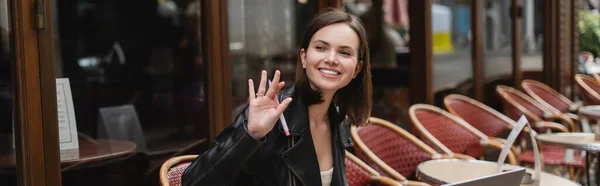 Happy woman in black jacket holding pen and waving hand in french outdoor cafe, banner — Stock Photo