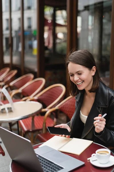 Cheerful freelancer in black jacket holding smartphone and pen near laptop in french outdoor cafe — Stock Photo