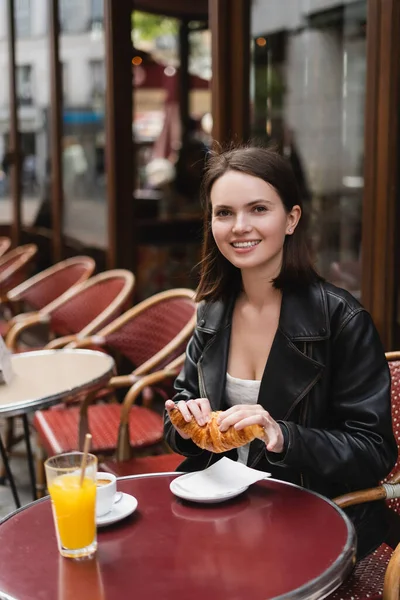 Felice donna che tiene croissant vicino a tazza di caffè e vetro di succo d'arancia in caffè all'aperto a Parigi — Foto stock