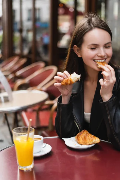 Mujer alegre comer croissant cerca de taza de café y vaso de jugo de naranja en la cafetería al aire libre en París - foto de stock
