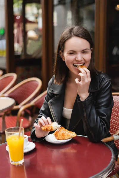 Mujer feliz comer croissant cerca de la taza de café y vaso de jugo de naranja en la cafetería al aire libre en París - foto de stock