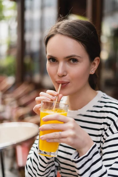Портрет женщины в полосатой рубашке с длинным рукавом, держащей стакан и пьющей апельсиновый сок в открытом кафе в Париже — стоковое фото