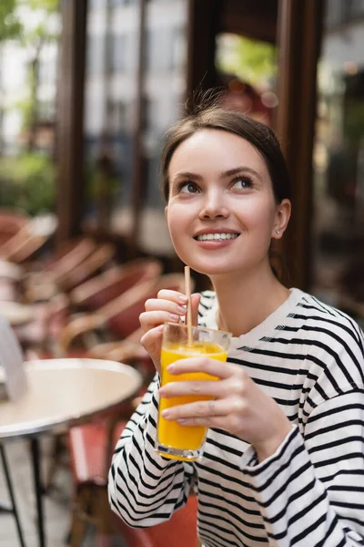 Femme gaie en chemise à manches longues rayée tenant un verre de jus d'orange frais dans un café en plein air à Paris — Photo de stock