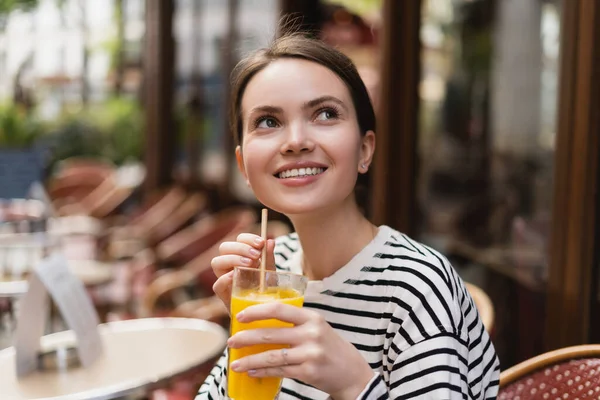 Mujer feliz con camisa de manga larga a rayas sosteniendo un vaso de jugo de naranja fresco en la cafetería al aire libre en París - foto de stock