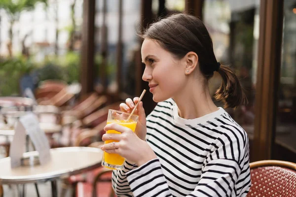 Vista lateral de la joven sonriente en camisa de manga larga a rayas sosteniendo un vaso de jugo de naranja fresco en la cafetería al aire libre en París - foto de stock
