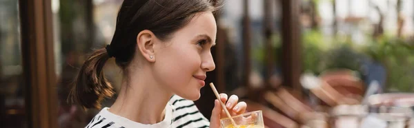 Vista laterale della giovane donna sorridente che tiene il bicchiere di succo d'arancia fresco nel caffè all'aperto a Parigi, banner — Foto stock