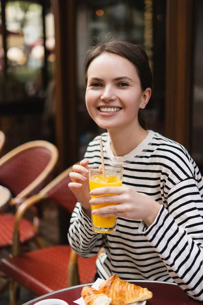 Радісна молода жінка в смугастій сорочці з довгим рукавом тримає склянку свіжого апельсинового соку у відкритому кафе в Парижі — стокове фото