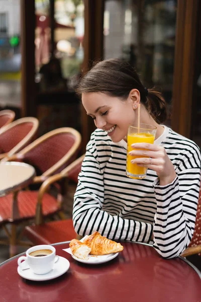 Щаслива жінка в смугастій сорочці з довгим рукавом тримає склянку свіжого апельсинового соку і дивиться на круасан у відкритому кафе в Парижі — стокове фото
