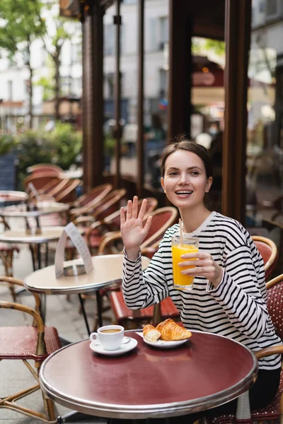 Щаслива жінка в смугастій сорочці з довгим рукавом тримає склянку апельсинового соку і махає рукою у відкритому кафе в Парижі — стокове фото