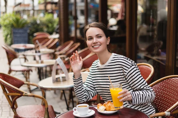 Mujer sonriente en camisa de manga larga a rayas sosteniendo un vaso de jugo de naranja y saludando la mano en la cafetería al aire libre en París - foto de stock