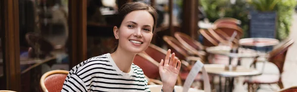 Mujer sonriente en camisa a rayas agitando la mano en la cafetería al aire libre en París, pancarta - foto de stock