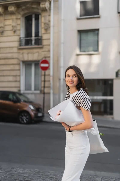 Fröhliche junge Frau in stilvollem Outfit mit in Papier gewickeltem Strauß auf der Straße in Paris — Stockfoto